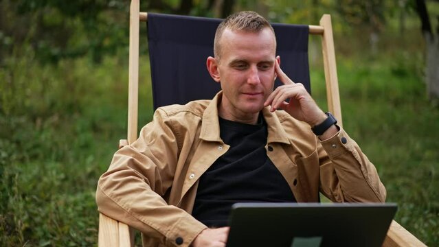 Pensive sitting in garden chair looks thoughtfully at his laptop screen. Freelancer working in nature has found some mistake and looks closer at computer.