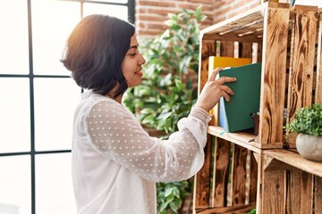 Young latin woman smiling confident holding book of shelving at home