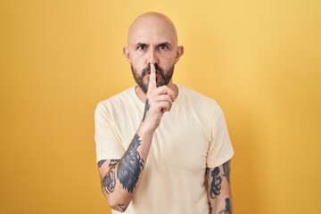 Hispanic man with tattoos standing over yellow background asking to be quiet with finger on lips. silence and secret concept.