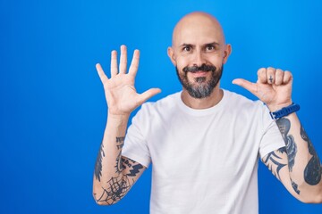 Hispanic man with tattoos standing over blue background showing and pointing up with fingers number six while smiling confident and happy.