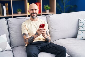 Young bald man using smartphone sitting on sofa at home