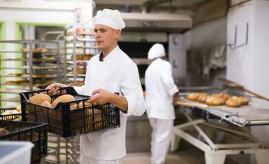 Baker in uniform holding crate with bread, industrial kitchen of bakery on background