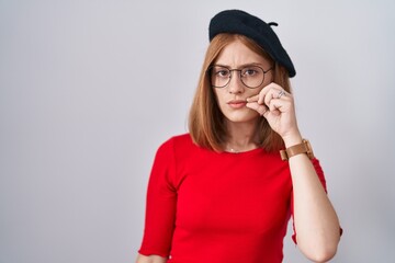 Young redhead woman standing wearing glasses and beret mouth and lips shut as zip with fingers....