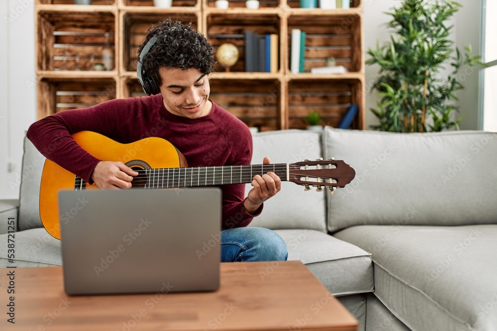 Sticker young hispanic man having online classical guitar lesson sitting on the sofa at home.