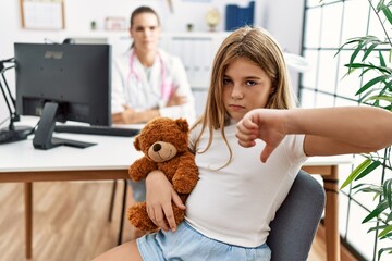 Blonde little girl at pediatrician clinic with female doctor with angry face, negative sign showing dislike with thumbs down, rejection concept