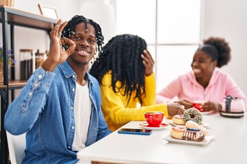 Group of three young black people sitting on a table having coffee doing ok sign with fingers, smiling friendly gesturing excellent symbol