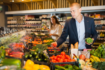 Obraz premium Young confident man carefully selects fresh vegetables on the counter in the supermarket