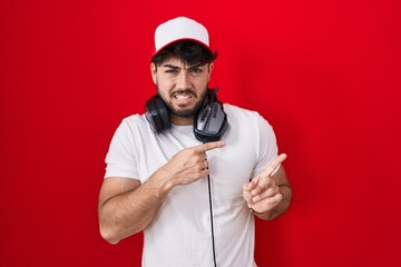 Hispanic man with beard wearing gamer hat and headphones pointing aside worried and nervous with...