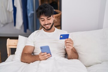 Young hispanic man using smartphone and credit card sitting on bed at bedroom