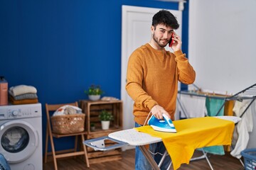 Young hispanic man talking on the smartphone ironing clothes at laundry room