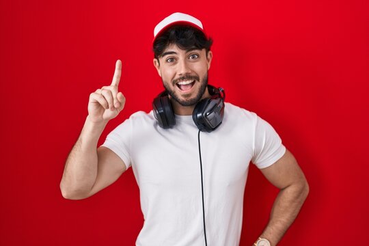 Hispanic man with beard wearing gamer hat and headphones pointing finger up with successful idea. exited and happy. number one.