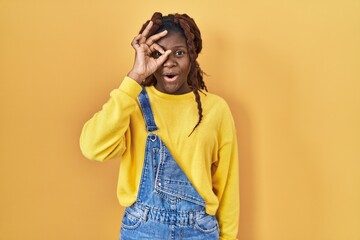 African woman standing over yellow background doing ok gesture shocked with surprised face, eye looking through fingers. unbelieving expression.