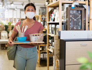 Asian woman customer in face mask choosing tableware and kitchen accessories at household goods...