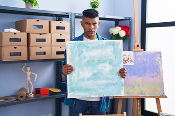Young hispanic man holding canvas at artist studio skeptic and nervous, frowning upset because of...