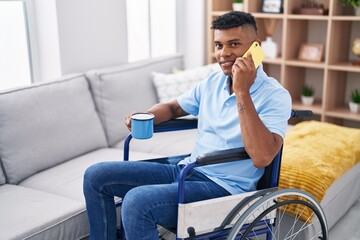 Young latin man talking on smartphone drinking coffee sitting on wheelchair at home