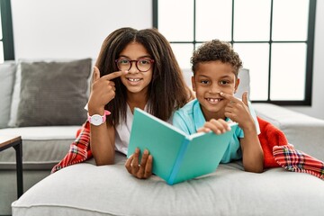 Two siblings lying on the sofa reading a book pointing with hand finger to face and nose, smiling...