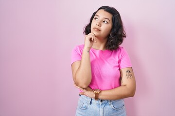 Young hispanic woman standing over pink background with hand on chin thinking about question, pensive expression. smiling with thoughtful face. doubt concept.