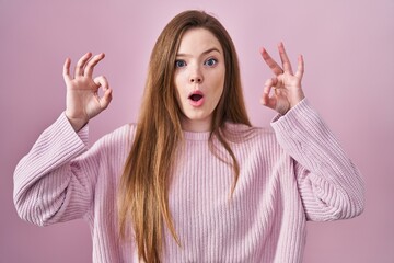 Young caucasian woman standing over pink background looking surprised and shocked doing ok approval symbol with fingers. crazy expression