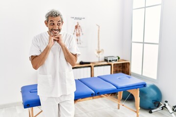 Obraz premium Middle age hispanic therapist man working at pain recovery clinic laughing nervous and excited with hands on chin looking to the side