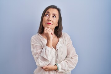 Middle age hispanic woman standing over blue background with hand on chin thinking about question,...