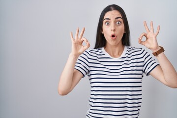 Young brunette woman wearing striped t shirt looking surprised and shocked doing ok approval symbol with fingers. crazy expression