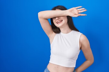 Fototapeta na wymiar Young caucasian woman standing over blue background covering eyes with arm smiling cheerful and funny. blind concept.
