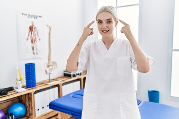 Young caucasian woman working at pain recovery clinic smiling pointing to head with both hands finger, great idea or thought, good memory