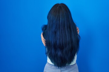 Naklejka premium Young modern girl with blue hair standing over blue background hugging oneself happy and positive from backwards. self love and self care