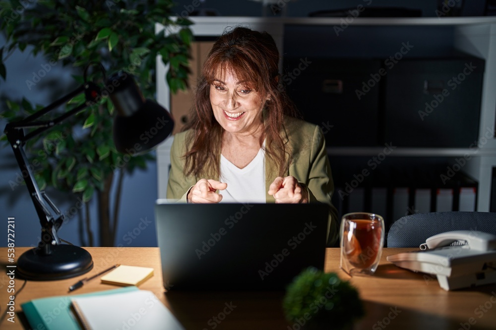 Poster middle age hispanic woman working using computer laptop at night pointing fingers to camera with hap