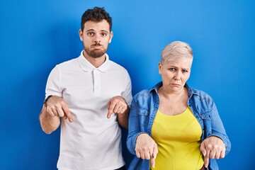 Young brazilian mother and son standing over blue background pointing down looking sad and upset,...