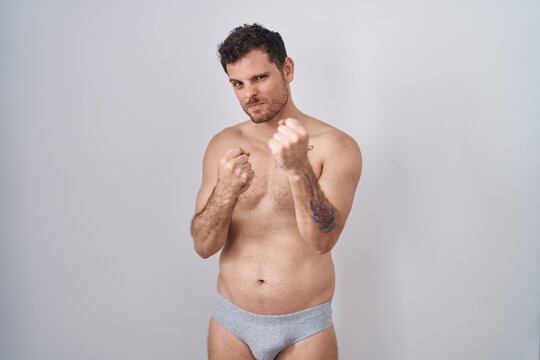 Young hispanic man standing shirtless wearing underware ready to fight with fist defense gesture, angry and upset face, afraid of problem