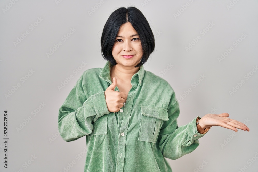 Poster Young asian woman standing over white background showing palm hand and doing ok gesture with thumbs up, smiling happy and cheerful