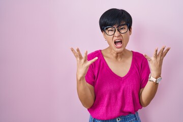 Obraz na płótnie Canvas Young asian woman with short hair standing over pink background crazy and mad shouting and yelling with aggressive expression and arms raised. frustration concept.