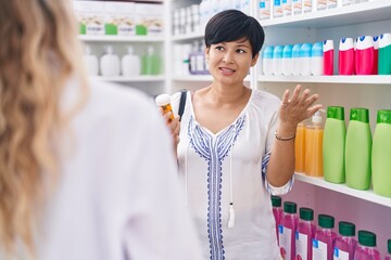 Middle age chinese woman customer holding pills speaking with pharmacist at pharmacy