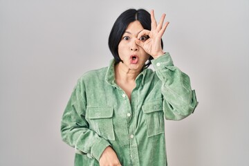 Young asian woman standing over white background doing ok gesture shocked with surprised face, eye looking through fingers. unbelieving expression.