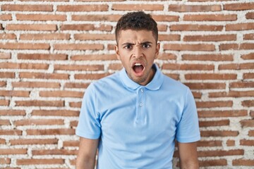 Brazilian young man standing over brick wall in shock face, looking skeptical and sarcastic, surprised with open mouth