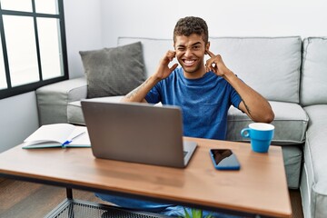 Young handsome hispanic man using laptop sitting on the floor covering ears with fingers with annoyed expression for the noise of loud music. deaf concept.