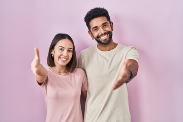 Obraz premium Young hispanic couple together over pink background smiling friendly offering handshake as greeting and welcoming. successful business.