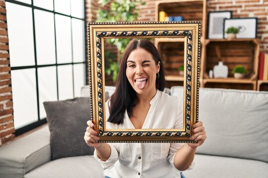 Young hispanic woman holding empty frame sticking tongue out happy with funny expression.