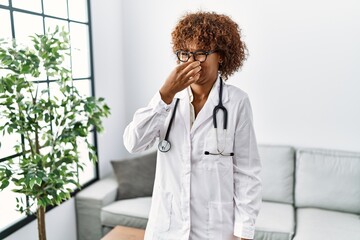 Young african american woman wearing doctor uniform and stethoscope smelling something stinky and...