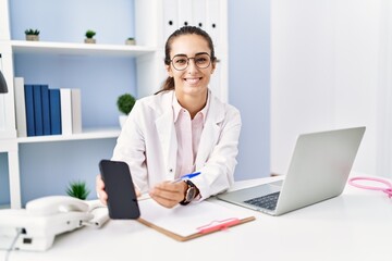 Young hispanic woman wearing doctor uniform showing smartphone screen at clinic