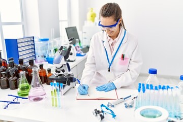 Fototapeta premium Young hispanic woman wearing scientist uniform writing on notebook using microscope at laboratory