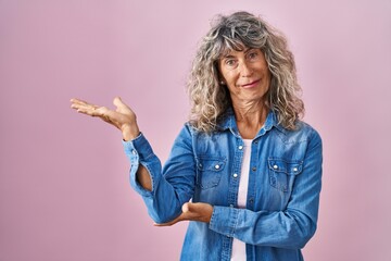 Middle age woman standing over pink background smiling cheerful presenting and pointing with palm of hand looking at the camera.
