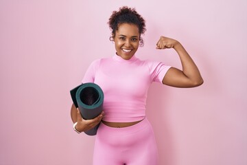 Young hispanic woman with curly hair holding yoga mat over pink background strong person showing arm muscle, confident and proud of power