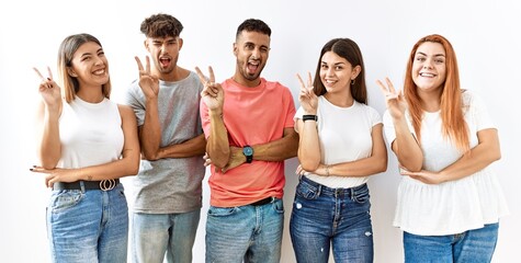 Group of young friends standing together over isolated background smiling with happy face winking at the camera doing victory sign with fingers. number two.