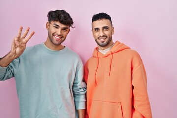 Young hispanic gay couple standing over pink background showing and pointing up with fingers number three while smiling confident and happy.