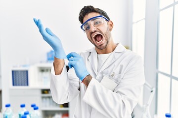 Young hispanic man working at scientist laboratory putting gloves on celebrating crazy and amazed for success with open eyes screaming excited.