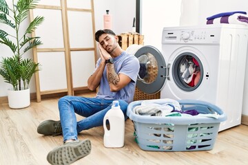 Young hispanic man putting dirty laundry into washing machine sleeping tired dreaming and posing with hands together while smiling with closed eyes.