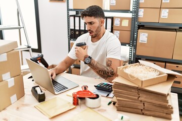 Young hispanic man working at small business ecommerce with laptop in shock face, looking skeptical and sarcastic, surprised with open mouth