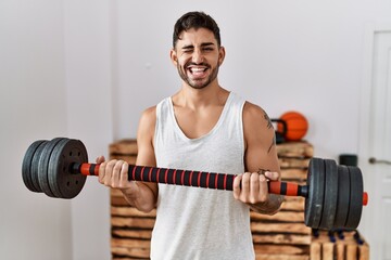 Young hispanic man training with weighs winking looking at the camera with sexy expression,...